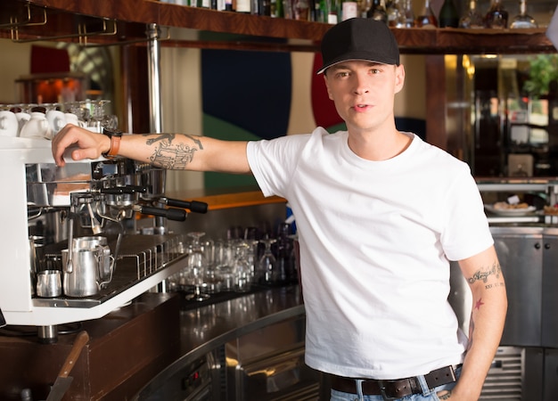 Joven tatuado barista serio de pie junto a la máquina de café de grado comercial de alta resistencia.