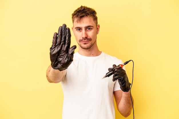 Joven tatuador caucásico aislado de fondo amarillo de pie con la mano extendida mostrando la señal de alto, impidiéndote.