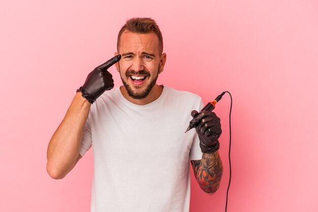 Joven tatuador caucásico aislado sobre fondo rosa mostrando un gesto de decepción con el dedo índice.