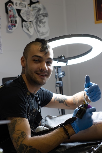 Joven tatuador realizando el gesto OK mientras tatúa en su estudio de tatuajes