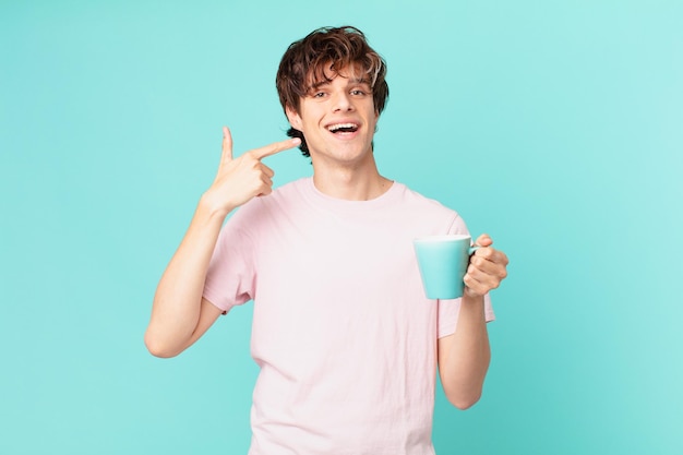 Joven con una taza de café sonriendo con confianza apuntando a su propia sonrisa amplia