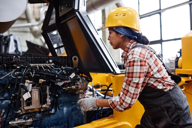 Joven técnica femenina en overoles y casco de seguridad revisando el motor de la máquina