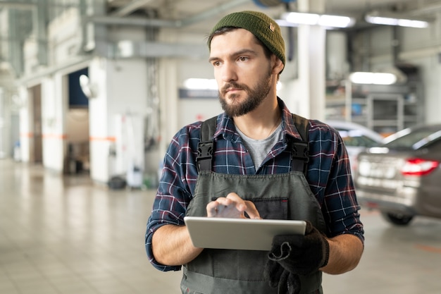 Joven técnico barbudo de servicio de automóvil contemporáneo tocando la pantalla de la tableta digital mientras mira a través de datos en línea sobre nuevos modelos