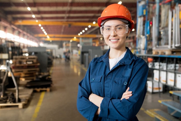 Foto joven técnico femenino sonriente en uniforme azul, anteojos protectores y casco trabajando en la fábrica moderna