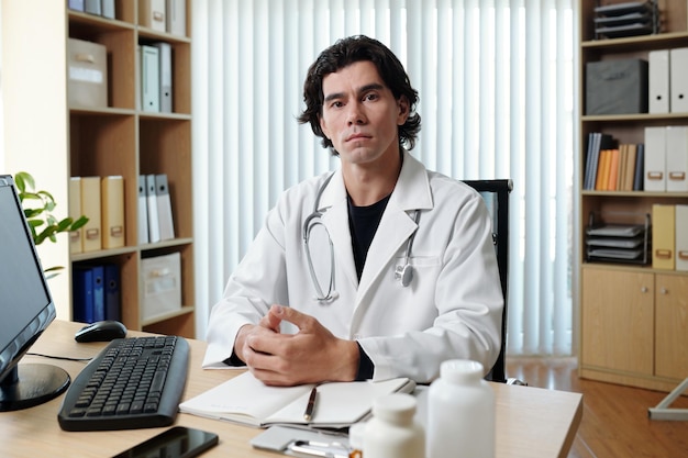 Joven teledoc masculino serio en bata de laboratorio sentado en el lugar de trabajo