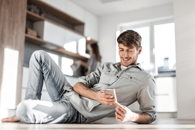 Joven con teléfono inteligente sentado en el piso de la cocina