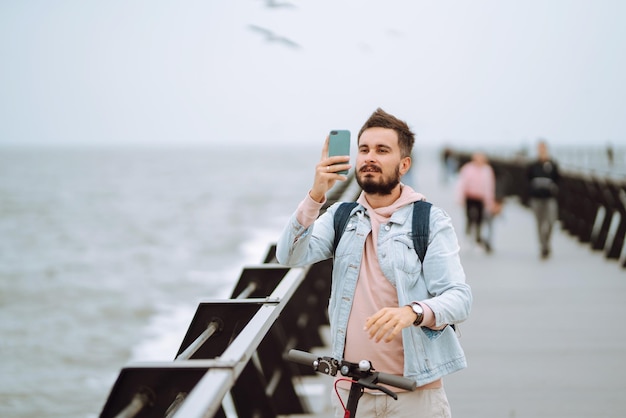 Joven con teléfono montando scooter eléctrico en un muelle cerca del mar Concepto de transporte ecológico