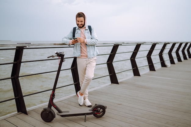 Joven con teléfono montando scooter eléctrico en un muelle cerca del mar Concepto de transporte ecológico