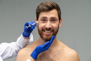 Foto joven temeroso de los procedimientos de belleza y cirugía plástica con una inyección rejuvenecedora en la frente por un médico con guantes aislados de fondo gris