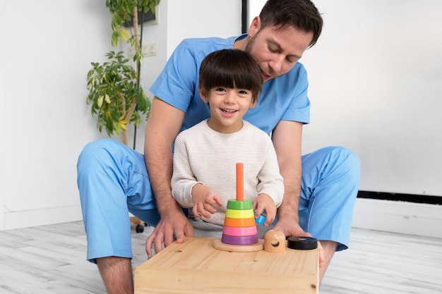 Joven terapeuta ayudando a un niño lindo que tiene parálisis cerebral jugando con un juguete en desarrollo en el centro de rehabilitación ...