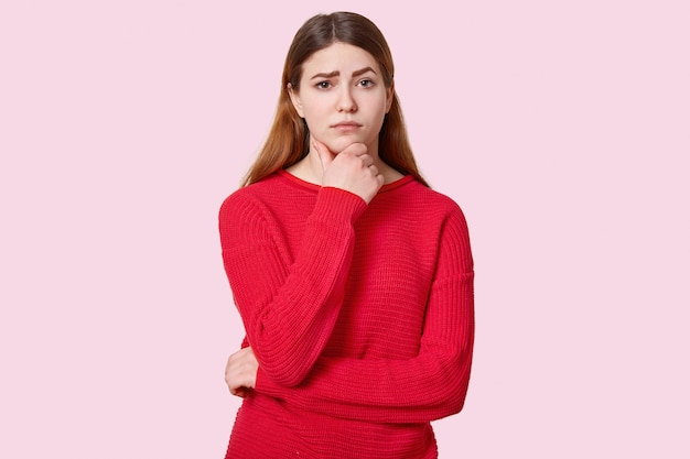 la joven tiene las manos parcialmente cruzadas sobre el pecho, mira con expresión triste, levanta las cejas oscuras, usa un atuendo rojo, se pone de color rosa. Triste modelo femenino