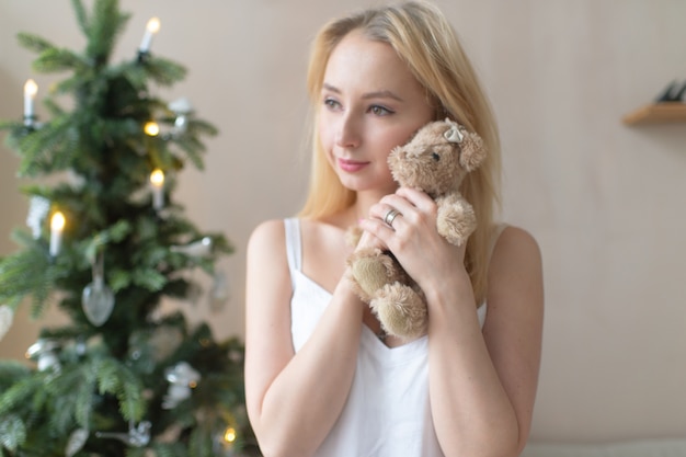 Joven tierna en camisón abraza oso de juguete con árbol de navidad