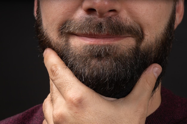 El joven toca la barba gruesa negra recortada con la mano después de los procedimientos de cuidado de la piel y el cabello en la barbería