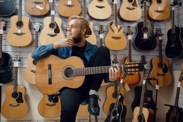 Joven toca la guitarra acústica en la tienda de música. Surtido en la tienda de instrumentos musicales, músico masculino comprando equipos