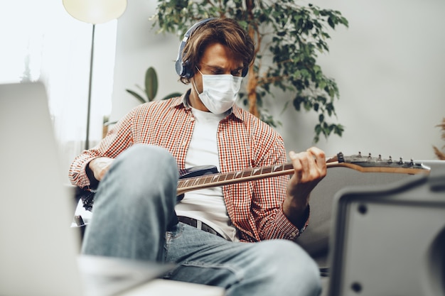 Joven toca la guitarra en casa con máscara médica. Concepto de estilo de vida de cuarentena de coronavirus