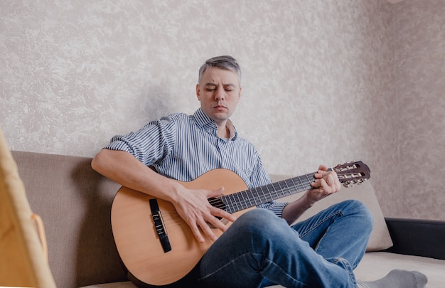 Un joven toca la guitarra sentado en un sofá en una luminosa sala de estar. Un instrumento musical para el concepto de ocio o pasatiempo. entretenimiento en el hogar