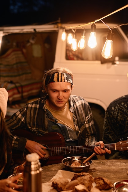 Joven tocando la guitarra para sus amigos mientras están sentados a la mesa durante el campamento en el bosque