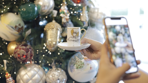 Joven tomando foto com seu telefone movil a una taza con un fondo de arbol navideno