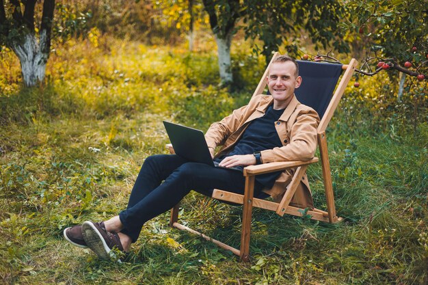 Un joven trabaja en una computadora portátil al aire libre Un joven autónomo descansa en el bosque Trabajo remoto recreación activa en el verano Concepto de personas de turismo hombre sentado en una silla al aire libre