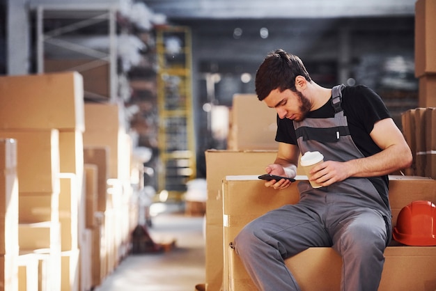 Foto joven trabajador de almacenamiento tiene un descanso, sentado en las cajas con el teléfono y una taza de bebida en las manos.