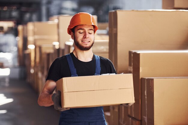 Joven trabajador de almacenamiento en uniforme y casco lleva caja en las manos.