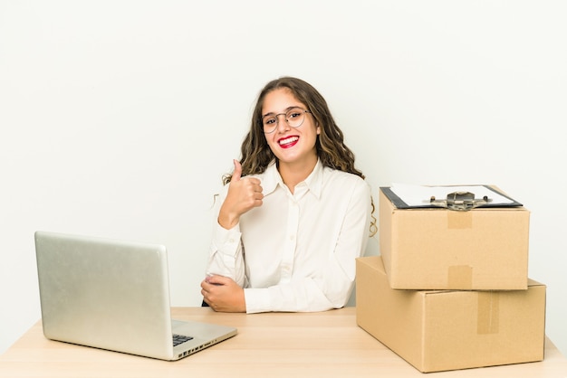 Joven trabajador caucásico de una empresa de paquetería aislada sonriendo y levantando el pulgar hacia arriba