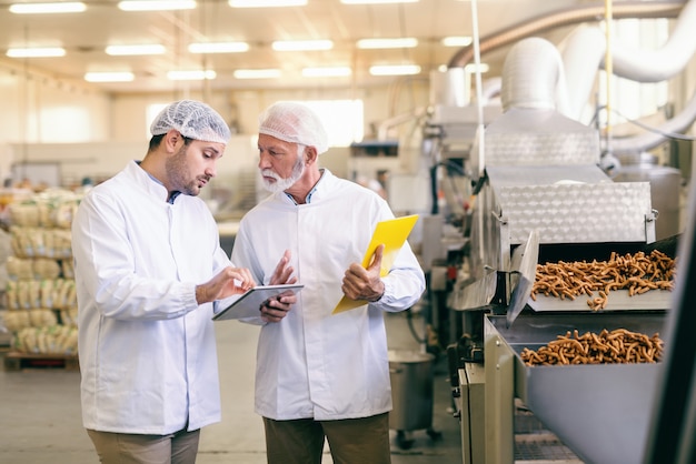 Joven trabajador caucásico que muestra a su colega mayor estadística en tableta mientras está de pie en la fábrica de alimentos.