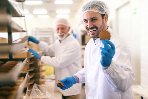 Foto joven trabajador caucásico sonriente mostrando la galleta mientras está de pie en la fábrica de alimentos.