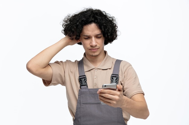 Joven trabajador de la construcción descontento con uniforme sosteniendo y mirando el teléfono móvil manteniendo la mano en la cabeza aislada en el fondo blanco