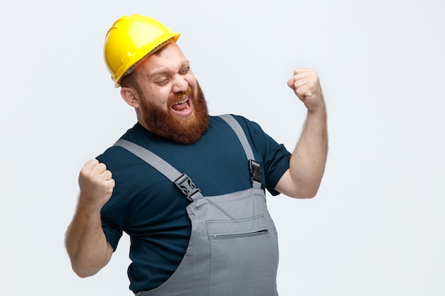 Joven trabajador de la construcción enojado con casco de seguridad y uniforme mirando al lado manteniendo los puños en el aire aislado en el fondo blanco