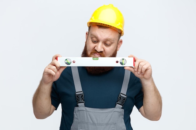 Un joven trabajador de la construcción inseguro que usa casco de seguridad y sostiene uniforme y mira el nivel de burbuja aislado de fondo blanco