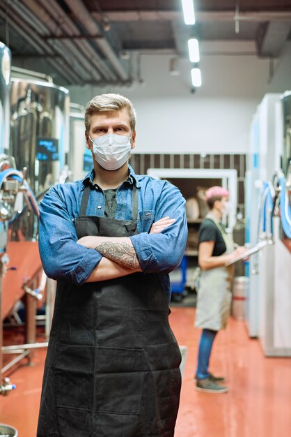 Joven trabajador de la fábrica de producción de cerveza en ropa de trabajo y máscara protectora de pie contra su colega reescribiendo datos desde el panel de control