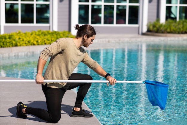 Joven trabajador limpiador profesional limpieza piscina con red de pala al aire libre.