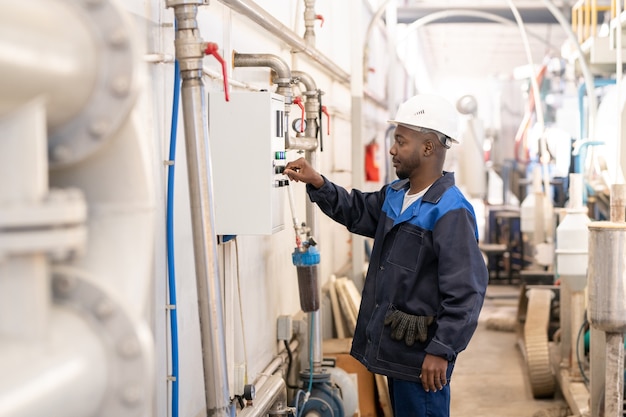 Joven trabajador masculino africano en monos y casco de pie delante de una enorme máquina industrial y girando la perilla en el panel de control