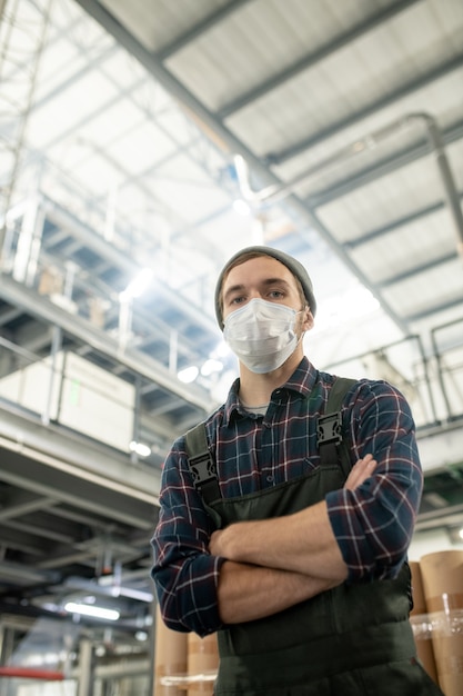 Joven trabajador masculino contemporáneo de gran fábrica en ropa de trabajo y máscara protectora de pie con los brazos cruzados delante de la cámara