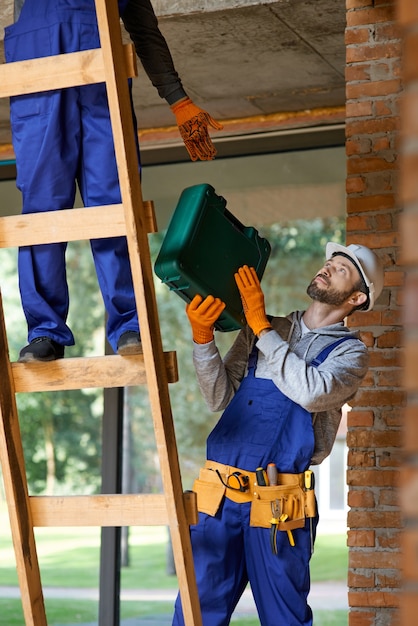 Foto joven trabajador en monos y casco dando caja de herramientas a su colega en la escalera mientras trabajaba en el sitio de construcción de la cabaña