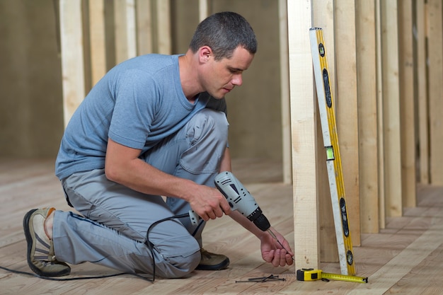 El joven trabajador profesional utiliza un nivel y un destornillador para instalar el marco de madera para paredes futuras.