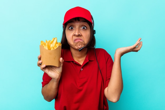 Joven trabajador de restaurante de comida rápida de mujer de raza mixta sosteniendo papas fritas aisladas sobre fondo azul se encoge de hombros y abre los ojos confundidos.