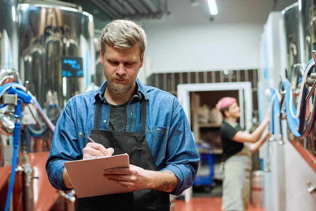 Joven trabajador de sexo masculino barbudo de la fábrica de producción de cerveza tomando notas mientras está de pie contra la cervecera trabajando por una gran máquina de procesamiento