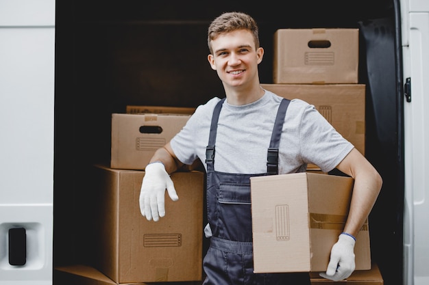 Un joven trabajador sonriente guapo con uniforme está de pie junto a la camioneta llena de cajas con una caja en sus manos. Mudanza de casa, servicio de mudanza.