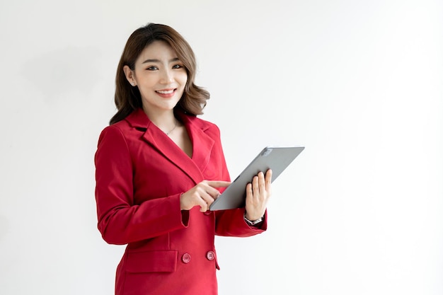 Joven trabajadora asiática de la empresa con traje rojo sonriendo y sosteniendo una tableta digital de pie sobre fondo blanco