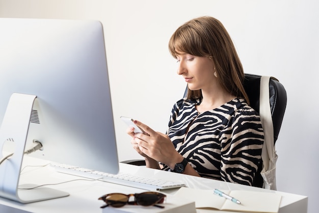 Joven trabajadora en el escritorio en la oficina moderna y minimalista. Retrato, de, mujer de negocios, utilizar, teléfono móvil, delante de, computadora de escritorio