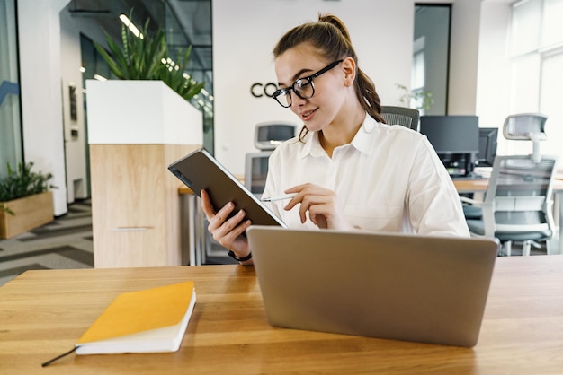 Una joven trabajadora examina atentamente su tableta, un bloc de notas y una computadora portátil a la mano