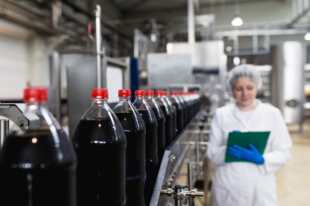 Foto joven trabajadora feliz en la fábrica de embotellado comprobando las botellas de jugo antes del envío. control de calidad de inspección.