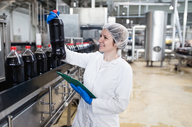 Joven trabajadora feliz en la fábrica de embotellado comprobando las botellas de jugo antes del envío. Control de calidad de inspección.