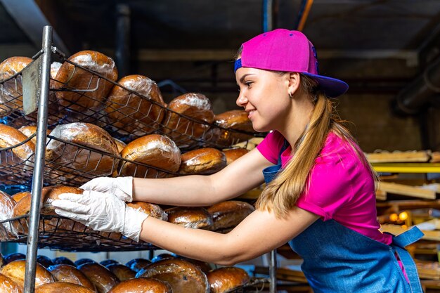 Joven trabajadora feliz en paños estériles sosteniendo muffins recién horneados en hojalata dentro de la fábrica de producción de alimentos.