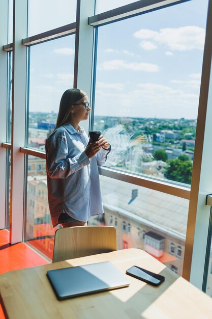 Una joven trabajadora independiente con anteojos está parada con un café cerca de una ventana grande y mirando la ciudad. Una chica con una camisa y pantalones cortos se encuentra en un café junto a la ventana.