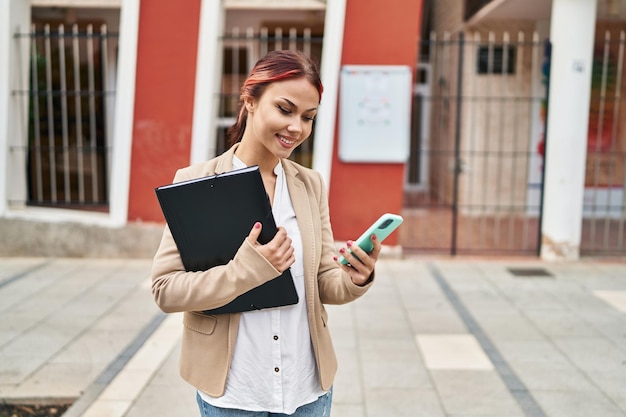 Joven trabajadora de negocios caucásica que usa un teléfono inteligente con carpeta en la calle