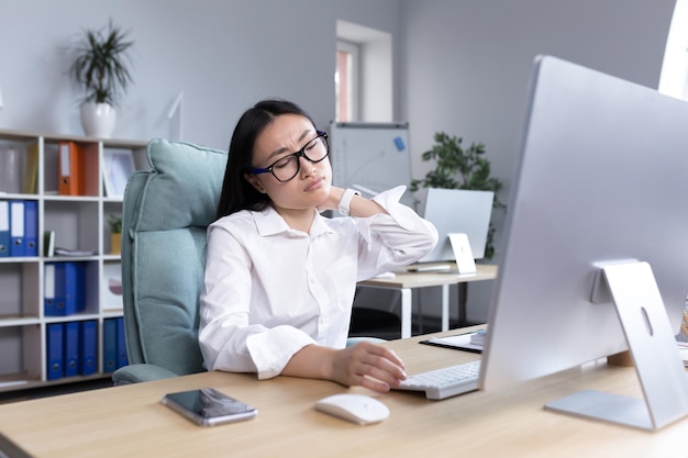 Una joven trabajadora de oficina asiática con gafas y una camisa blanca se sienta en un escritorio en una computadora cansada