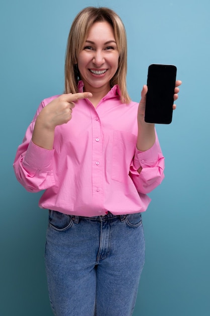 La joven trabajadora de oficina europea rubia con el pelo suelto en una blusa rosa muestra una maqueta de teléfono con